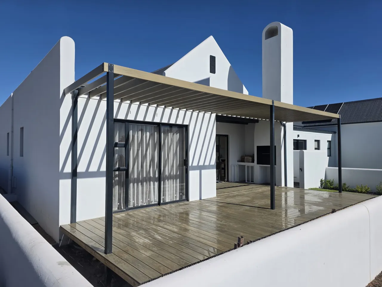 Timber deck under a slatted pergola on a white Cape-style house