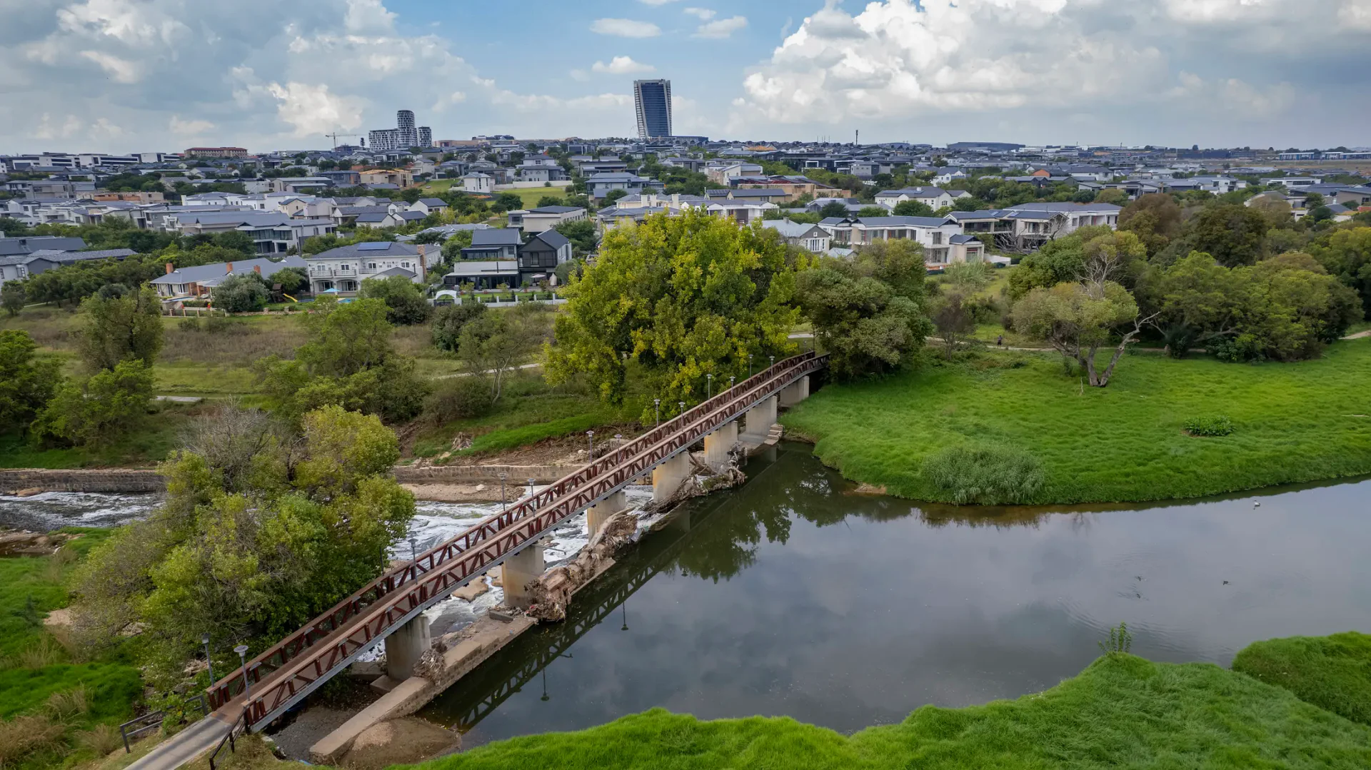 Aerial view of a red steel golf cart bridge crossing a river, city skyline beyond
